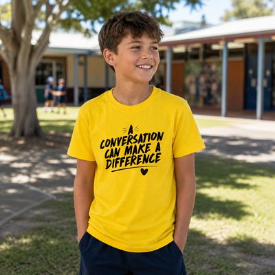 A smiling young boy wearing a bright yellow t-shirt with the black printed text 'A CONVERSATION CAN MAKE A DIFFERENCE' and dark navy shorts, standing outdoors in front of a school building.