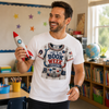 A man in a classroom setting wearing a white graphic t-shirt that reads 'THIS IS MY BOOK WEEK COSTUME' with a space astronaut design, while holding a toy rocket.