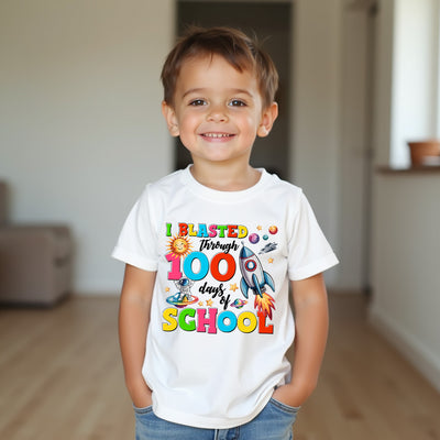 A young boy smiling in a bright indoor room, wearing a white graphic T-shirt that reads 'I BLASTED Through 100 days of SCHOOL' with colorful space-themed illustrations including a rocket, planets, and a sun.