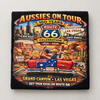 A black square fabric item, likely a sweatshirt or towel, featuring a colorful graphic print of an Australian-themed 'Aussies on Tour' Route 66 celebration. The graphic includes illustrations of people in a red convertible car on a desert road trip with stops in Chicago, Las Vegas, and Santa Monica, and text reading '100 Years Route 66 Celebration 1926 - 2026'.