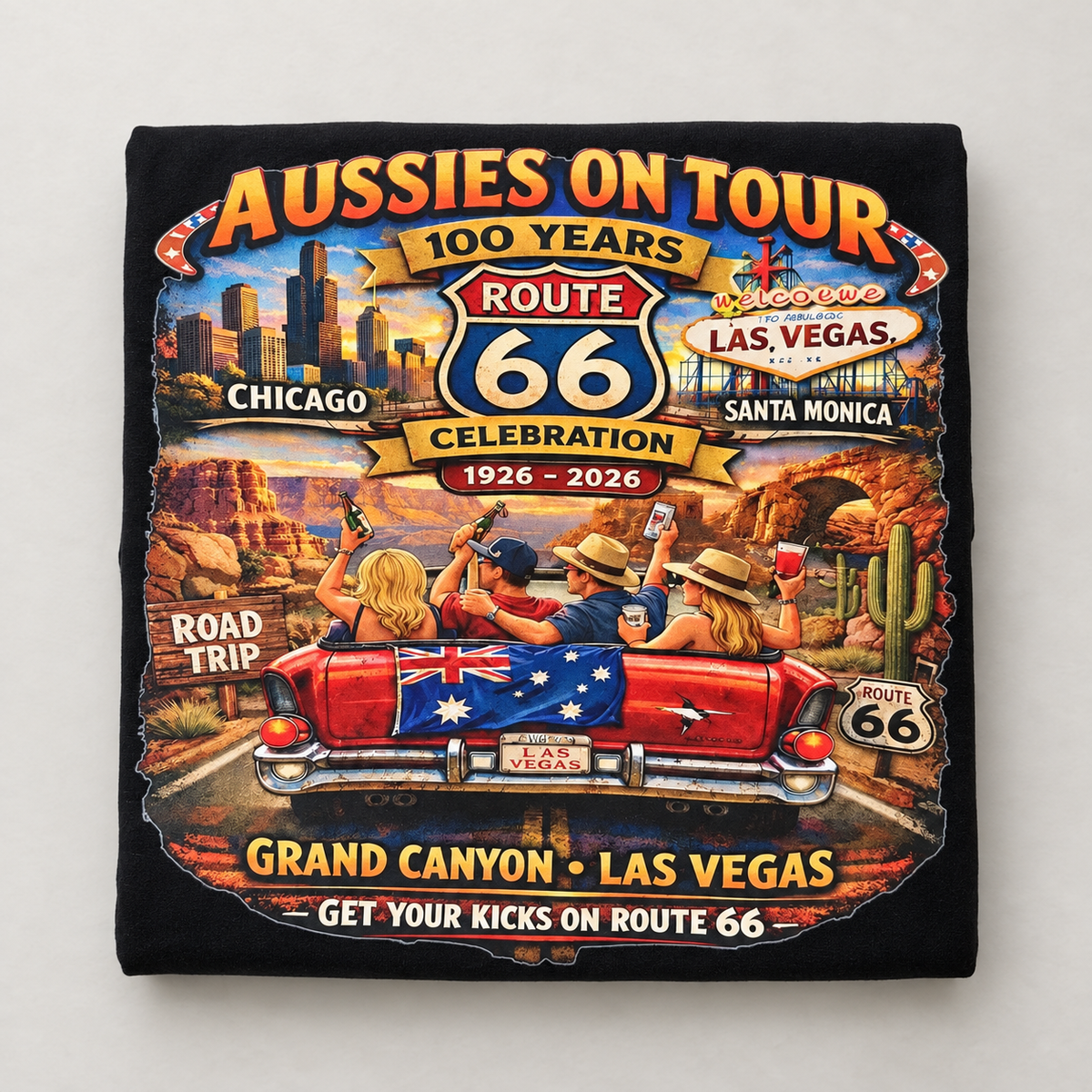 A black square fabric item, likely a sweatshirt or towel, featuring a colorful graphic print of an Australian-themed 'Aussies on Tour' Route 66 celebration. The graphic includes illustrations of people in a red convertible car on a desert road trip with stops in Chicago, Las Vegas, and Santa Monica, and text reading '100 Years Route 66 Celebration 1926 - 2026'.
