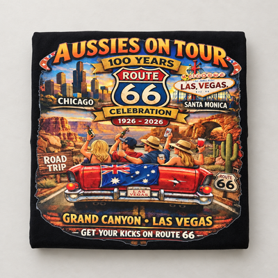 A black square fabric item, likely a sweatshirt or towel, featuring a colorful graphic print of an Australian-themed 'Aussies on Tour' Route 66 celebration. The graphic includes illustrations of people in a red convertible car on a desert road trip with stops in Chicago, Las Vegas, and Santa Monica, and text reading '100 Years Route 66 Celebration 1926 - 2026'.