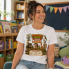 A woman wearing a white graphic t-shirt with an Australian Zoo Adventure in the Wild print, sitting in a classroom or library setting.