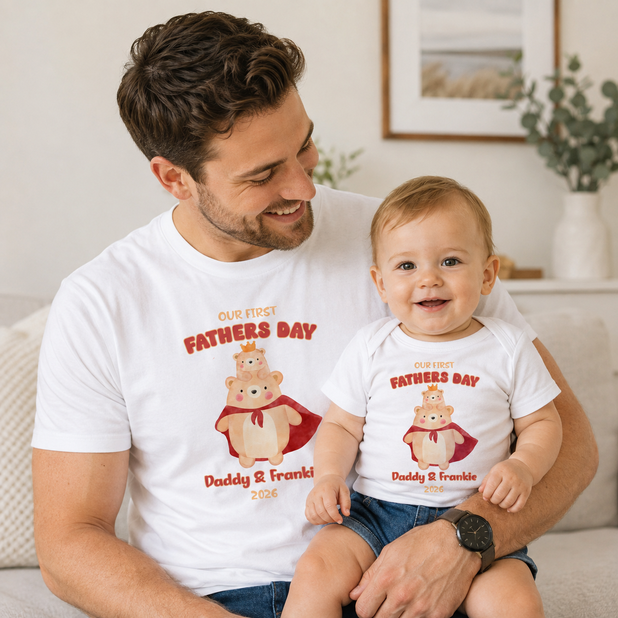 A man and a baby sitting together on a couch, both wearing matching white graphic t-shirts with a cartoon bear illustration and the text 'OUR FIRST FATHERS DAY Daddy & Frankie 2026'.