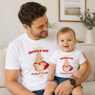 A man and a baby sitting together on a couch, both wearing matching white graphic t-shirts with a cartoon bear illustration and the text 'OUR FIRST FATHERS DAY Daddy & Frankie 2026'.