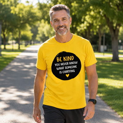 A smiling man walking on a sunny park path wearing a bright yellow t-shirt with a black circular graphic that reads 'BE KIND YOU NEVER KNOW WHAT SOMEONE IS CARRYING' in yellow and white text.