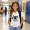 A young girl smiling in a school hallway, wearing a white graphic t-shirt with a black rainbow design and the words 'BETTER TOGETHER' on the front, paired with light blue jeans and a black backpack.