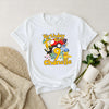 White t-shirt with colorful graphics and text on a light surface with flowers and a mug.
