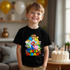Child wearing a black t-shirt with colorful graphics and text, standing in a room with balloons and a cake.