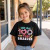 A young girl smiling while wearing a black graphic t-shirt that reads 'Little Miss 100 days SMARTER' with colorful donut and lollipop graphics, standing outdoors on a sidewalk.