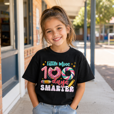 A young girl smiling while wearing a black graphic t-shirt that reads 'Little Miss 100 days SMARTER' with colorful donut and lollipop graphics, standing outdoors on a sidewalk.
