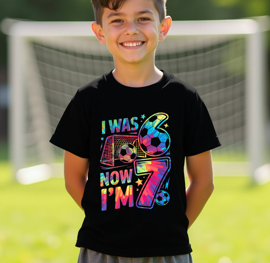 Child wearing a black t-shirt with colorful text 6 7 and numbers, standing in front of a soccer goal.