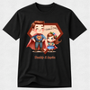 A black short-sleeve crewneck t-shirt with a custom cartoon graphic of a father and daughter dressed as superheroes, with 'Father's Day' and 'Daddy & Layla' text, displayed on a light gray textured background.
