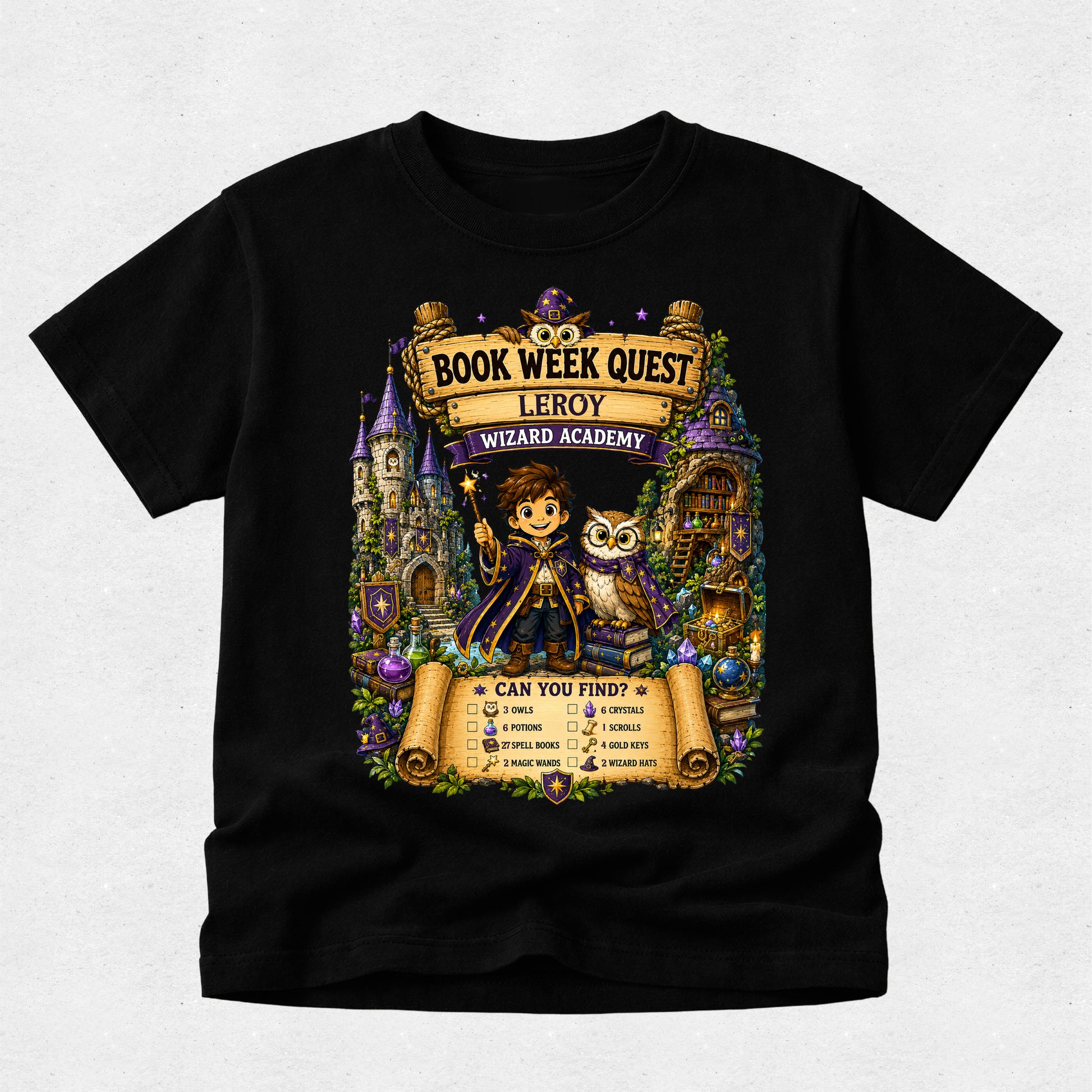 A black short-sleeve crewneck t-shirt featuring a large, colorful graphic print on the front. The graphic is themed around 'Book Week Quest' at the 'Leroy Wizard Academy' and shows an illustration of a young boy wizard holding a wand next to an owl, with scrolls, castles, and magical items in the background.