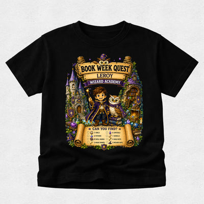 A black short-sleeve crewneck t-shirt featuring a large, colorful graphic print on the front. The graphic is themed around 'Book Week Quest' at the 'Leroy Wizard Academy' and shows an illustration of a young boy wizard holding a wand next to an owl, with scrolls, castles, and magical items in the background.