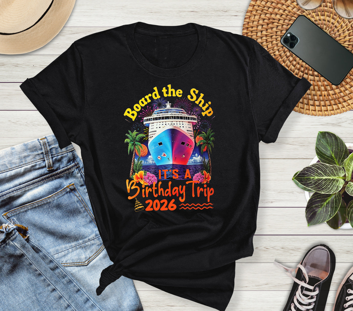 Black t-shirt with colorful ship graphic and text 'Board the Ship It's a Birthday Trip 2026' on a wooden surface with jeans and shoes.