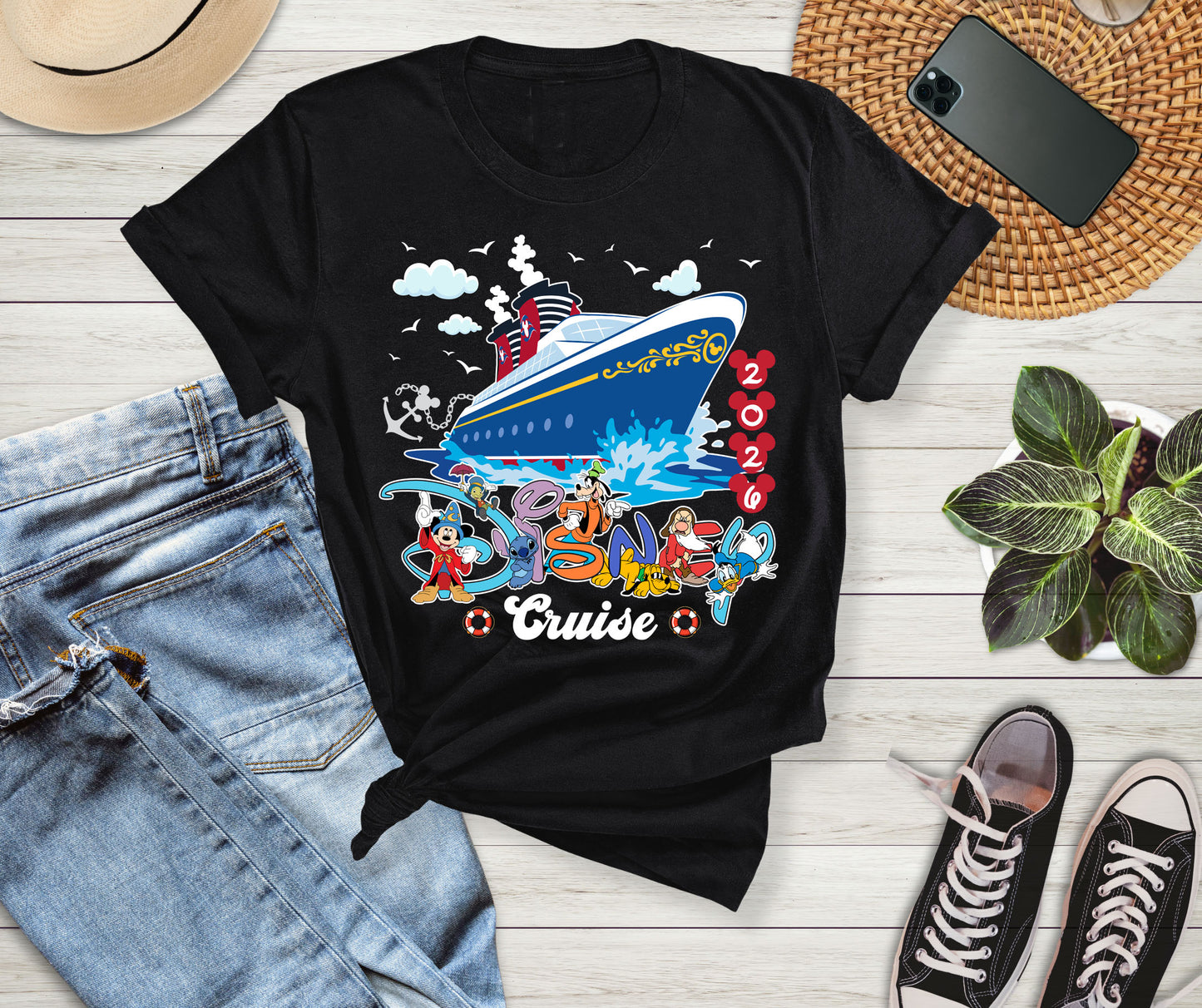 Black t-shirt with cruise ship graphic on a wooden surface with jeans, sneakers, and a phone.