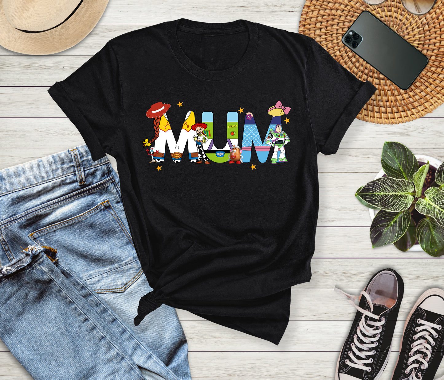 Black t-shirt with colorful 'MUM' design on a wooden surface with jeans, sneakers, and a phone.