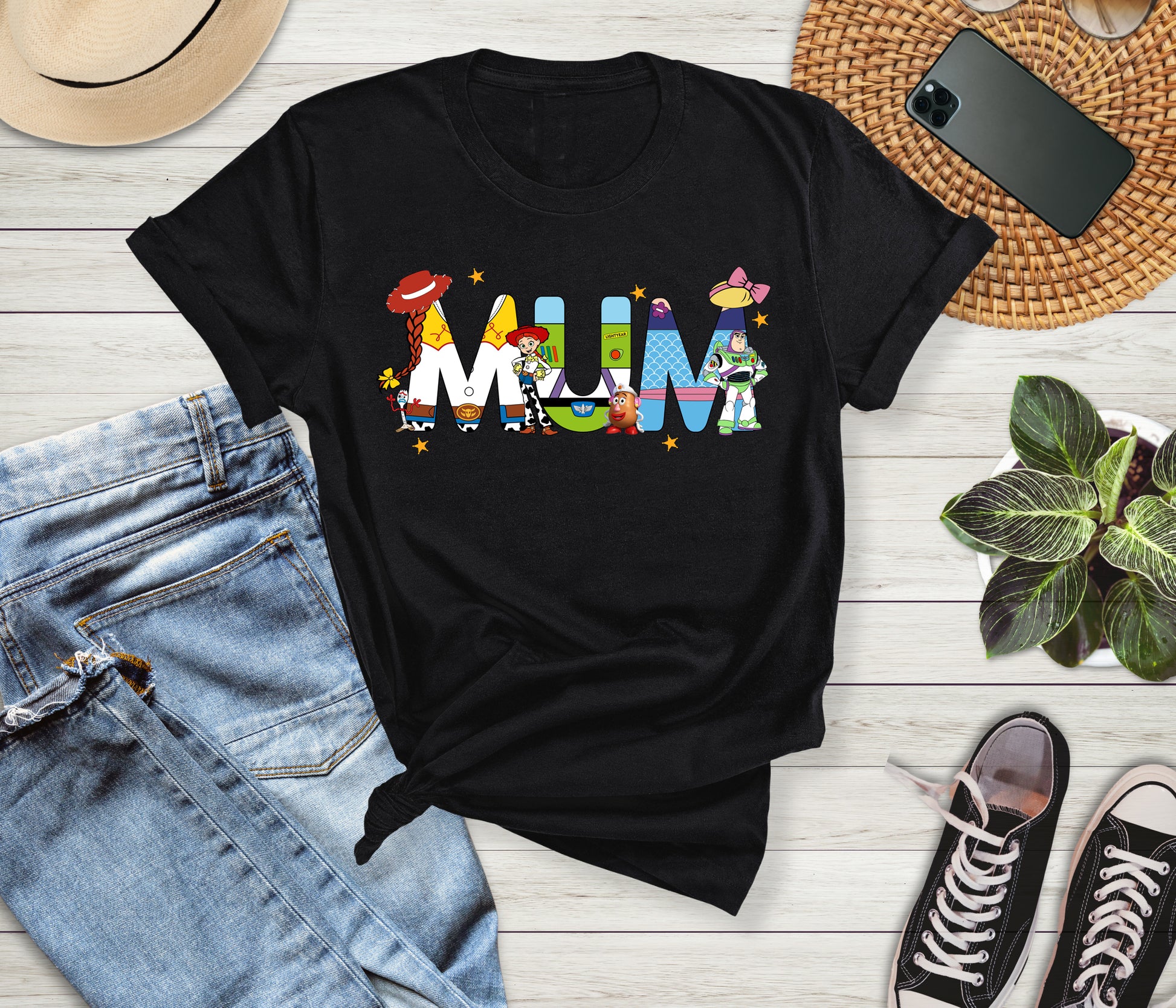 Black t-shirt with colorful 'MUM' design on a wooden surface with jeans, sneakers, and a phone.