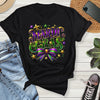 Black t-shirt with Mardi Gras design on a wooden surface with jeans, sneakers, and a phone.