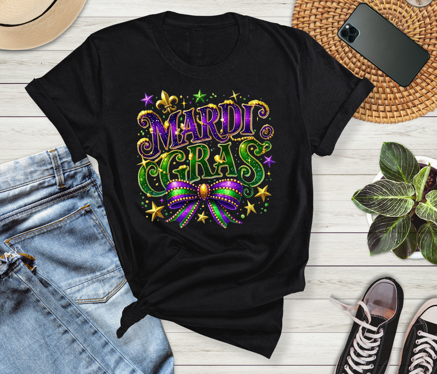 Black t-shirt with Mardi Gras design on a wooden surface with jeans, sneakers, and a phone.