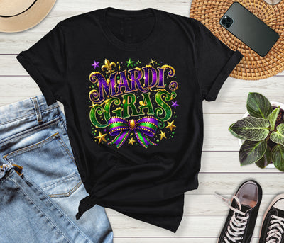 Black t-shirt with Mardi Gras design on a wooden surface with jeans, sneakers, and a phone.