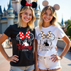 Two women wearing Disney-themed shirts with castle and bow designs in front of a castle.