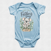 A light blue short-sleeve baby bodysuit with a Father's Day graphic on the front, featuring a koala illustration, eucalyptus leaves, and the text 'Our First Father's Day 2026'.
