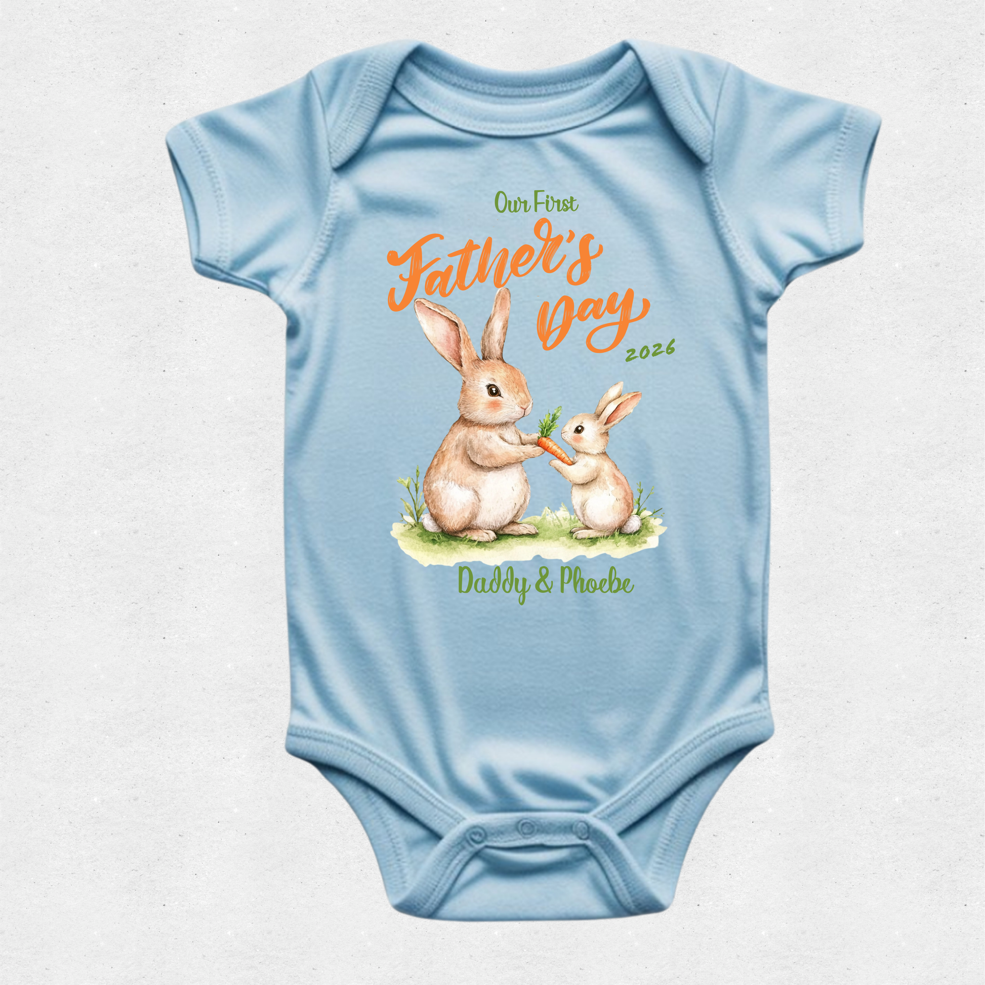 A light blue short-sleeve baby bodysuit with a custom Father's Day graphic. The graphic features an illustration of a mother rabbit and her baby, with orange text reading 'Our First Father's Day 2026' and green text reading 'Daddy & Me' at the bottom, displayed on a textured light gray background.