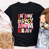 Black t-shirt with colorful text and cartoon characters, surrounded by white sneakers, a straw bag, denim shorts, and a hat.