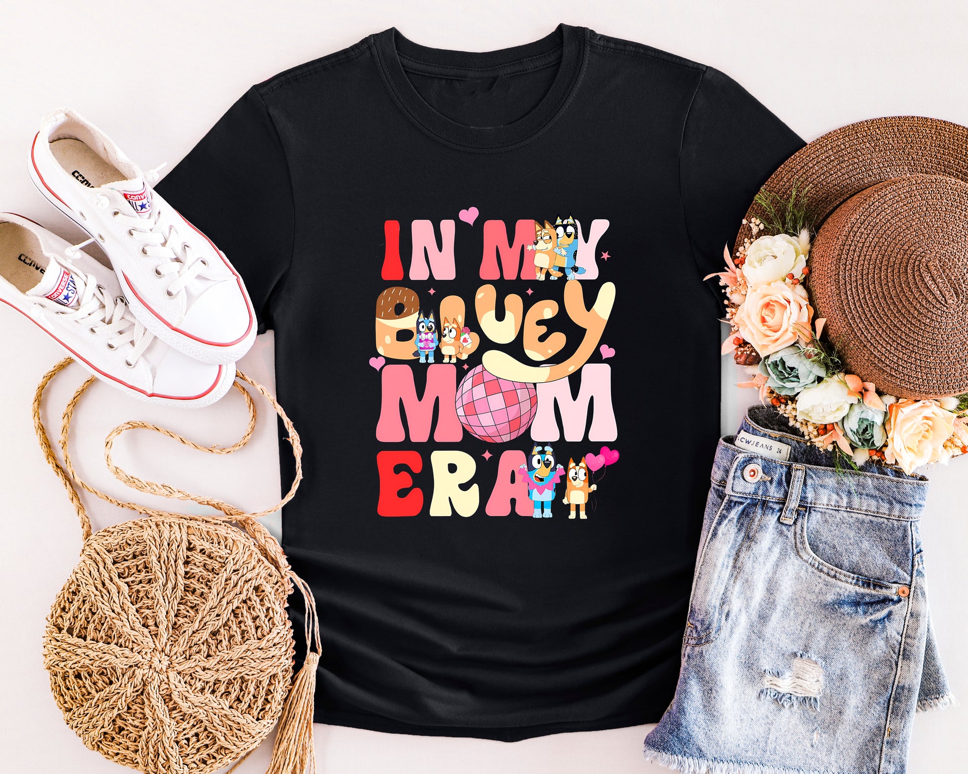 Black t-shirt with colorful text and cartoon characters, surrounded by white sneakers, a straw bag, denim shorts, and a hat.