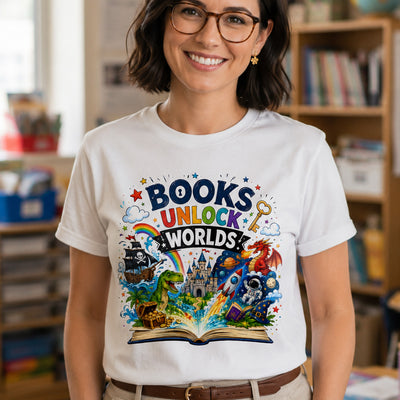 A smiling woman with glasses and short brown hair wears a white graphic T-shirt in a softly blurred indoor room. The shirt features a colorful printed design with the words 'BOOKS UNLOCK WORLDS' above an open book and fantasy/sci-fi illustrations including a rainbow, castle, dragon, rocket, and astronaut.