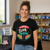 A smiling woman with her hair in a bun stands in a classroom or library setting wearing a black graphic t-shirt that reads 'IT'S BOOK WEEK' inside a colorful circular design. Behind her are bookshelves filled with books and bins.