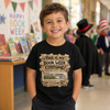 A smiling young boy stands in a bookstore wearing a black graphic t-shirt that reads 'THIS IS MY BOOK WEEK COSTUME! COSTUME LOADING.
