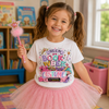 A young girl with brown pigtails smiles while wearing a white graphic T-shirt that says 'This is my Book Week Costume!' and a pink tutu skirt. She holds a pink sparkly wand in a brightly lit children's room with bookshelves in the background.