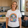 A smiling woman stands indoors wearing a white graphic T-shirt with a colorful book-themed illustration and the words 'IT'S BOOK WEEK!' on the front. The background shows a bright office or breakroom with a table, chairs, kitchen cabinets, and a sofa.