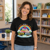 A smiling woman stands indoors wearing a black graphic T-shirt with a colorful book-themed design that reads 'IT'S BOOK WEEK!' and shows a rainbow, books, stars, and a cartoon character. The background shows a softly blurred classroom or library setting with bookshelves, a table, and plants.