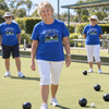 An older woman walking on a lawn bowling green wearing a blue River City Bowls Club t-shirt and white capris, with two other men in matching shirts standing in the background.