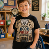 A smiling young boy stands in a classroom holding a magnifying glass, wearing a black graphic T-shirt that reads 'THIS IS MY BOOK WEEK COSTUME!' with a detective costume illustration, and blue jeans.