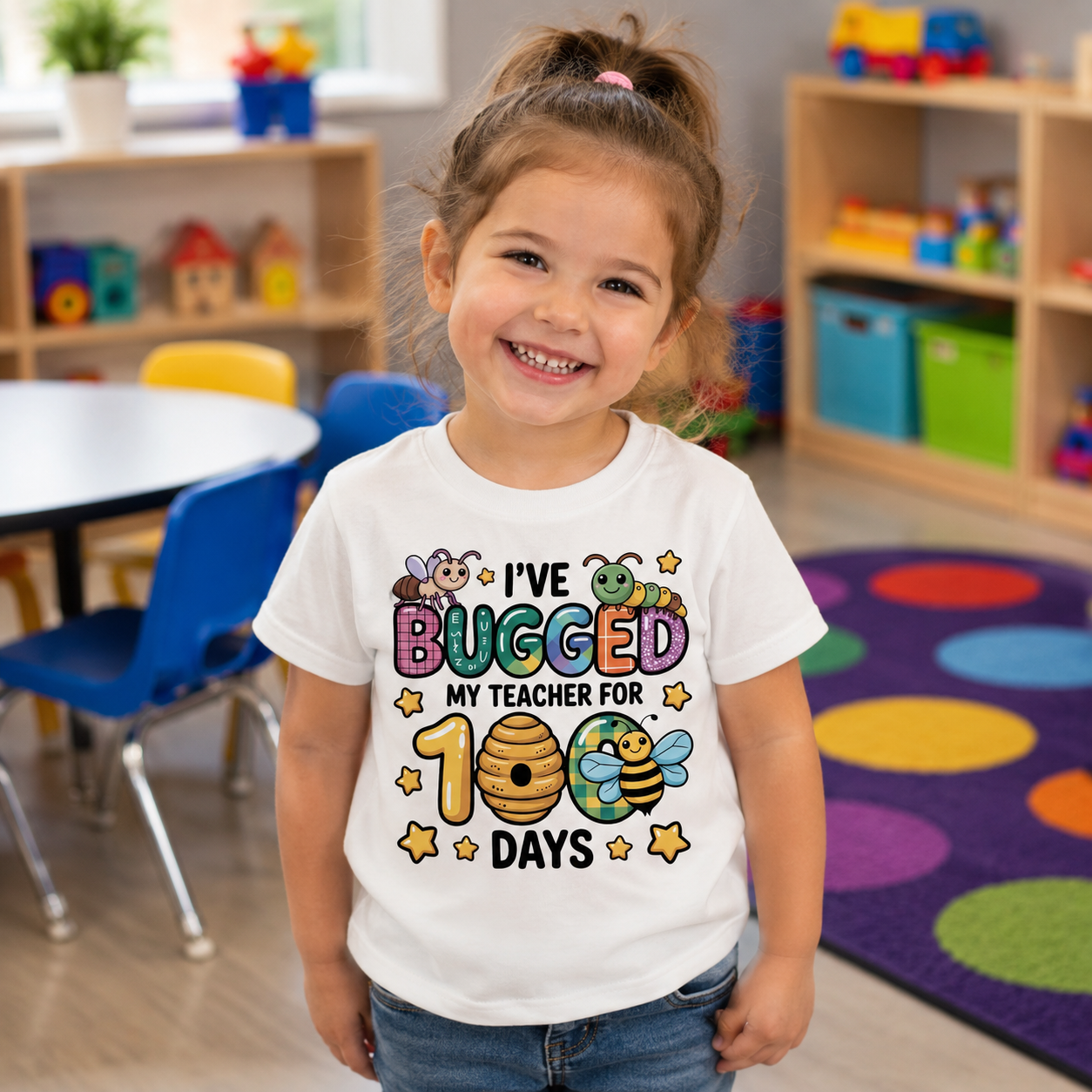A young girl smiling in a colorful children's classroom, wearing a white graphic t-shirt that reads 'I'VE BUGGED MY TEACHER FOR 100 DAYS' with cartoon bees and stars.