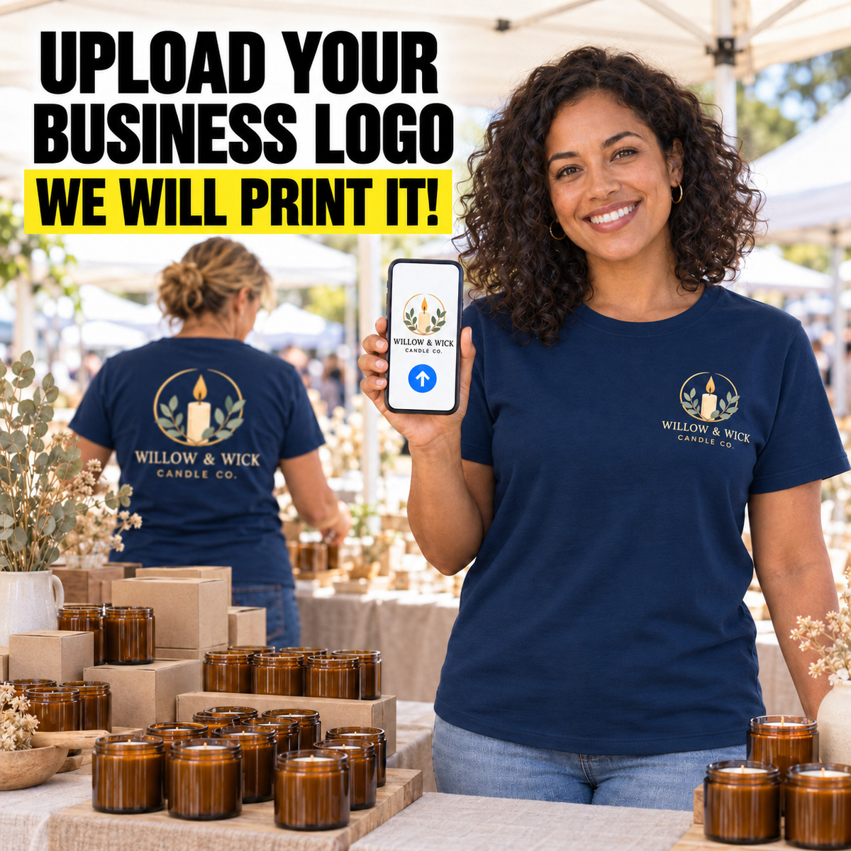 A smiling woman at an outdoor market wears a navy T-shirt with a printed 'Willow & Wick Candle Co.' logo on the chest and holds up a phone showing the same logo design.