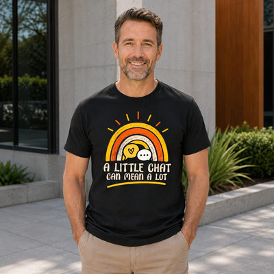 A smiling man wearing a black graphic t-shirt with a retro rainbow design and the text 'A LITTLE CHAT CAN MEAN A LOT', standing outdoors in front of a modern building.