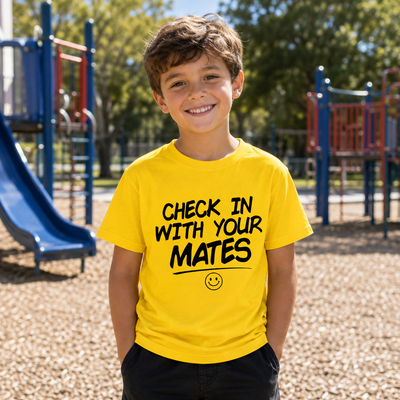 A young boy smiling in a bright yellow t-shirt with the text 'CHECK IN WITH YOUR MATES' and a smiley face graphic, standing in a playground with wood chips and blue play equipment in the background.