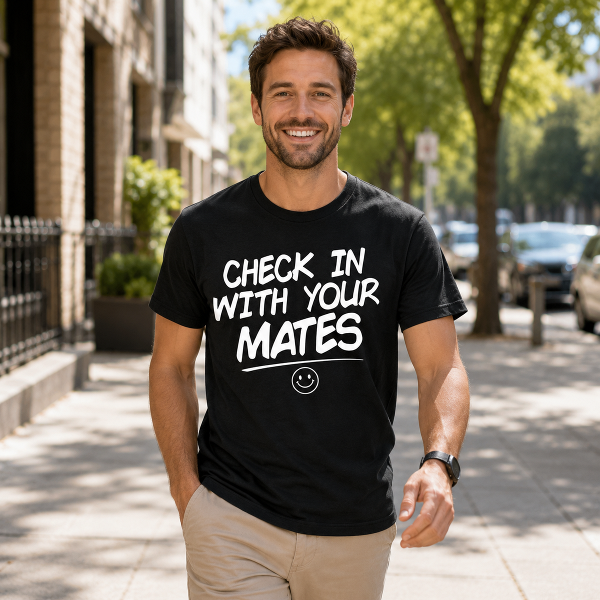 A smiling man standing on a sunny city sidewalk wearing a black graphic t-shirt with the white text 'CHECK IN WITH YOUR MATES' and a small smiley face underneath.