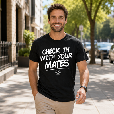 A smiling man standing on a sunny city sidewalk wearing a black graphic t-shirt with the white text 'CHECK IN WITH YOUR MATES' and a small smiley face underneath.