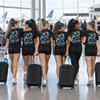 Six female cheerleaders walk away in an airport terminal wearing matching black t-shirts with a colorful graphic and the text 'VELOCITY CHEER 2026 SALT LAKE CITY'. They are pulling black rolling suitcases.