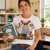 A woman smiling in a classroom setting, wearing a white graphic t-shirt with a 'Little Chef' cookbook and school uniform design that reads 'THIS IS MY BOOK WEEK COSTUME!'. She is holding a 'Little Chef' cookbook.