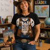 A smiling woman wearing a brown cowboy hat, a graphic t-shirt with a cowboy costume print, and blue jeans stands in a classroom setting holding a book titled 'The Cowboy and the Book Club'.