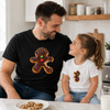 A man and a young girl sitting at a kitchen table, both wearing matching graphic t-shirts featuring a gingerbread cookie with colorful sprinkles. The man wears a black t-shirt, and the girl wears a white t-shirt.