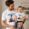 A smiling man and baby wearing matching white graphic T-shirts with a blue 'Our First Father's Day' print and a dinosaur illustration, posed indoors with a neutral background.